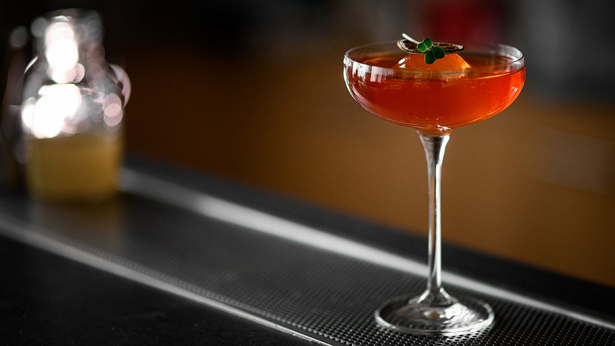 Stemmed glass filled with red brown cocktail garnished with citrus fruits and green leaves garnish.