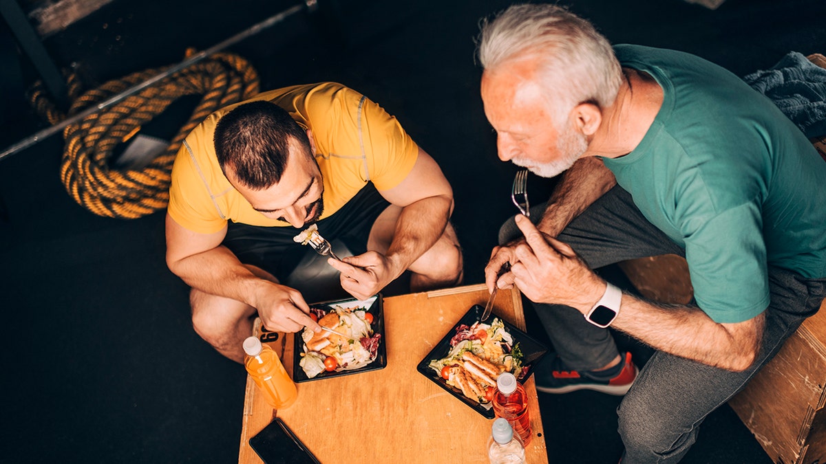 Active elderly sportsman eating a healthy protein chicken salad meal in a take-out box with his personal coach in a gym area of a gym.