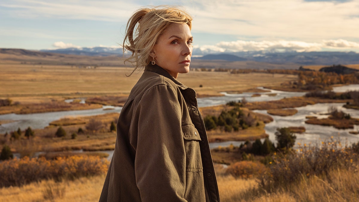 Michelle Pfeiffer standing on a hillside overlooking a wide valley with winding rivers and mountains in the distance.