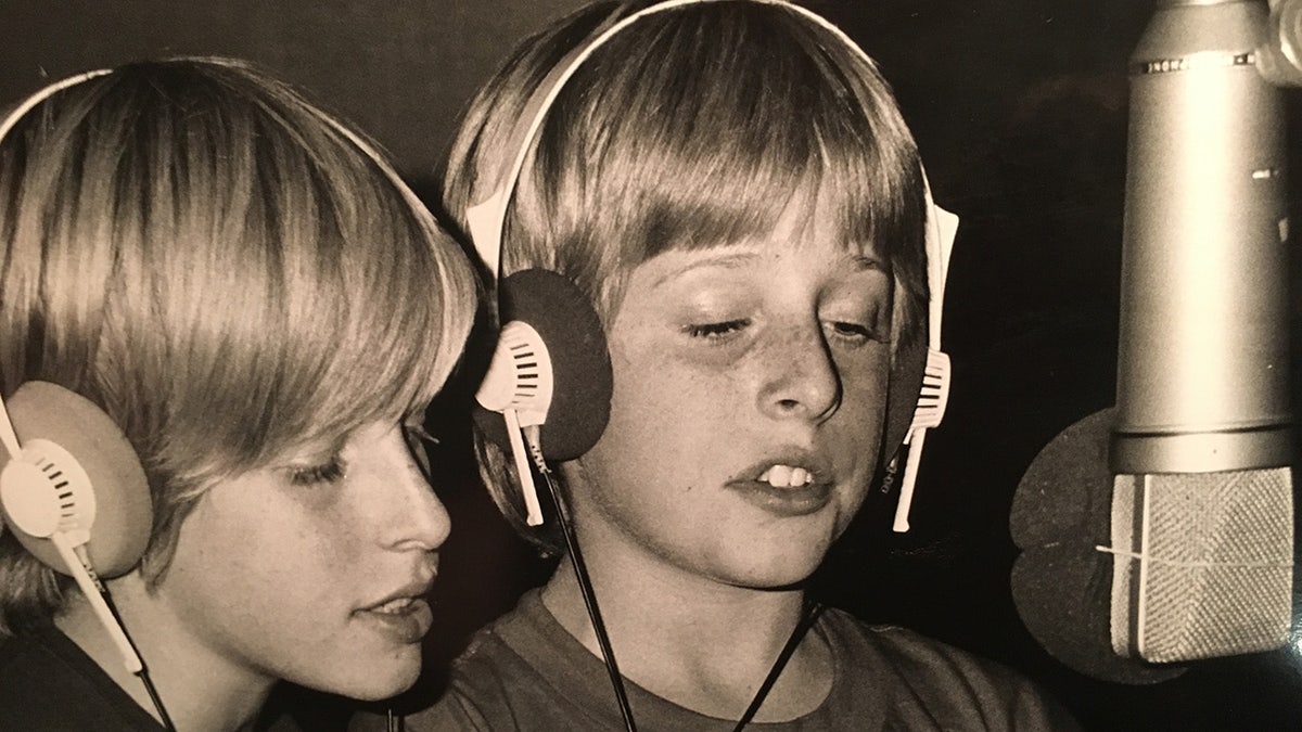 Gunnar and Matt Nelson wearing over-ear headphones record vocals together in a studio, standing close to a large microphone.
