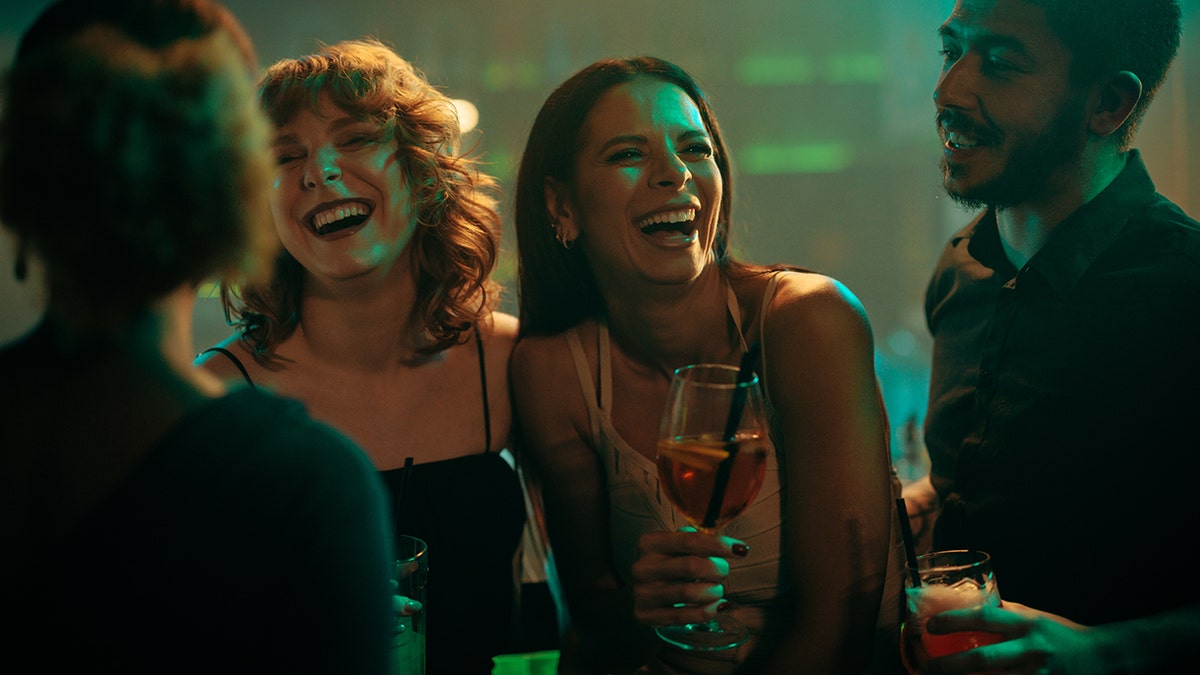 Due giovani donne che ridono del club mentre tengono i loro cocktail in piedi accanto a un bell'uomo con la barba.