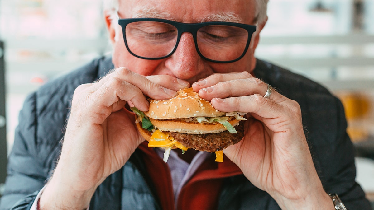 Older antheral with glasses eating a cheeseburger.