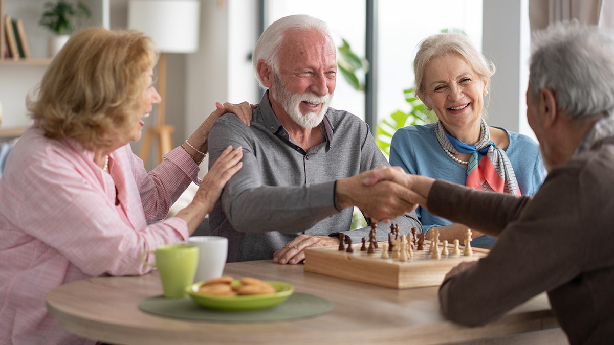 A group of senior friends joyfully engaging in a chess game, showcasing camaraderie and enjoyment in a warm, inviting environment filled with greenery.