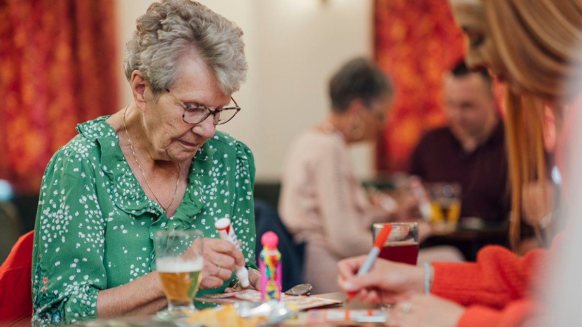 A senior woman sitting with her granddaughter in a social club in Newcastle upon Tyne, England. They are playing bingo together and marking numbers off their bingo card with a felt tip pen while looking down and concentrating.
