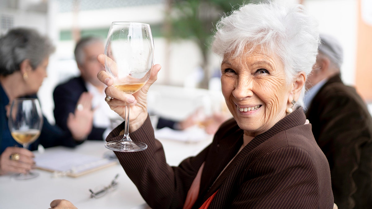 Portrait of smiling senior women holding a wine glass toward camera with friends behind her dining.