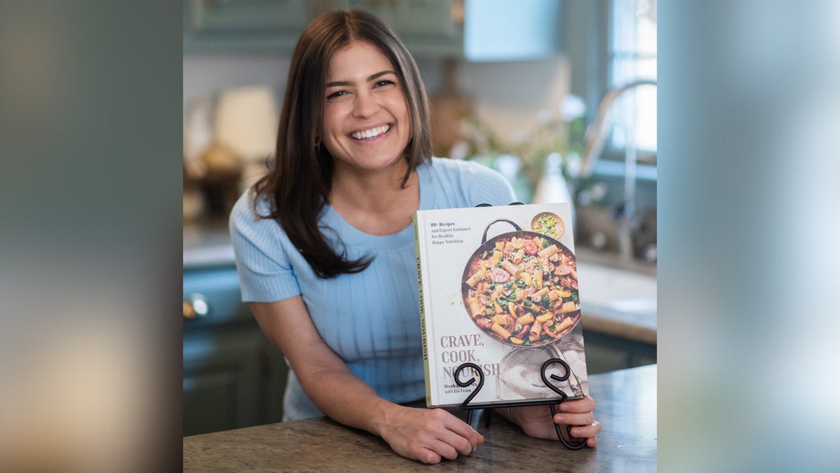 Registered dietitian Steph Grasso smiling in kitchen holding her new cookbook, "Crave, Cook, Nourish."
