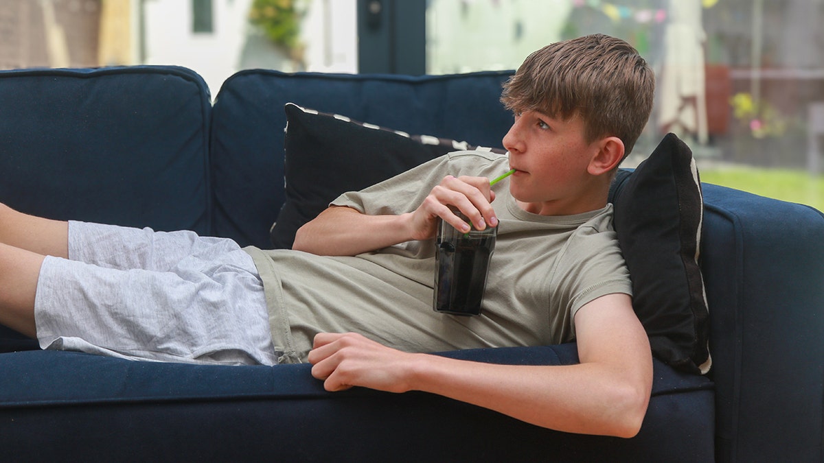 the boy is lying on the sofa drinking a soda