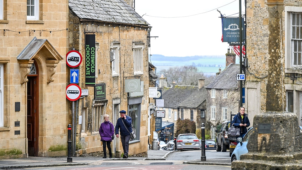 Historic Cotswold village street with stone buildings, pedestrians walking past local shops, and parked cars in a scenic English countryside setting.