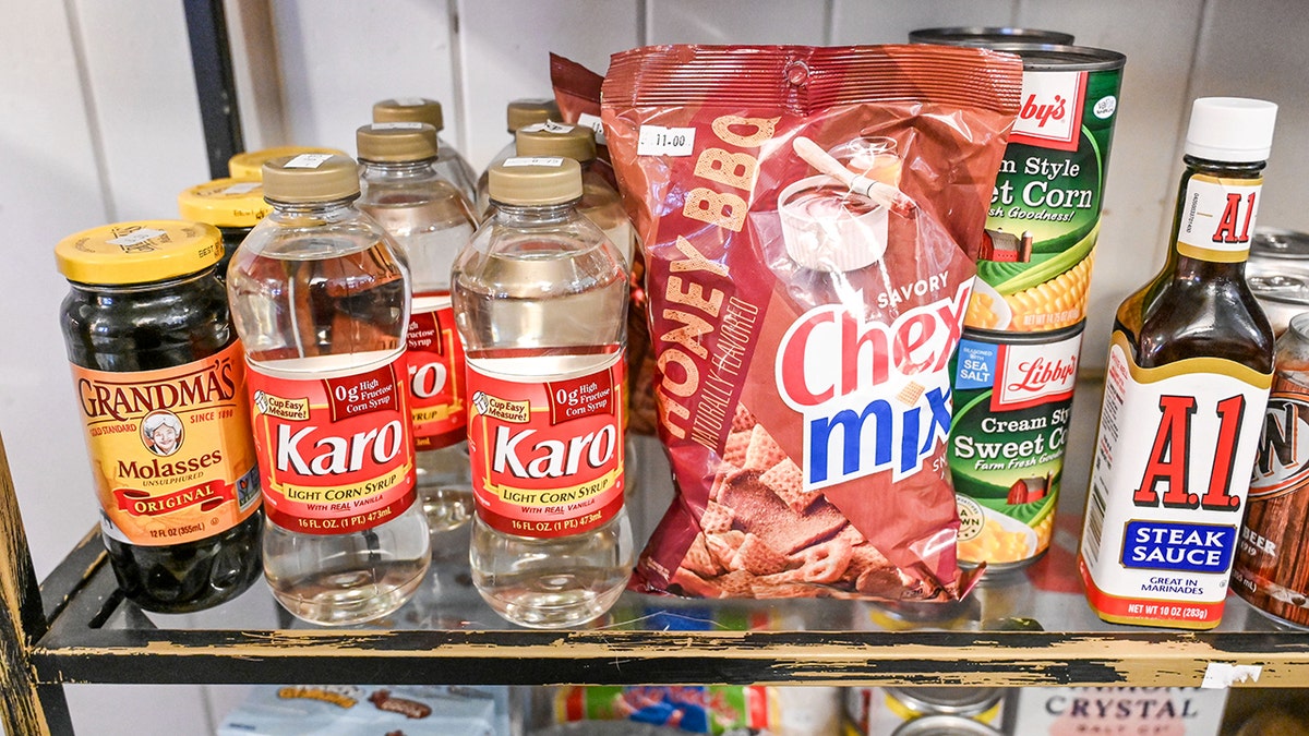 Assorted pantry staples including Karo corn syrup, Grandma’s molasses, Chex Mix, canned corn, and A1 steak sauce on a kitchen shelf.