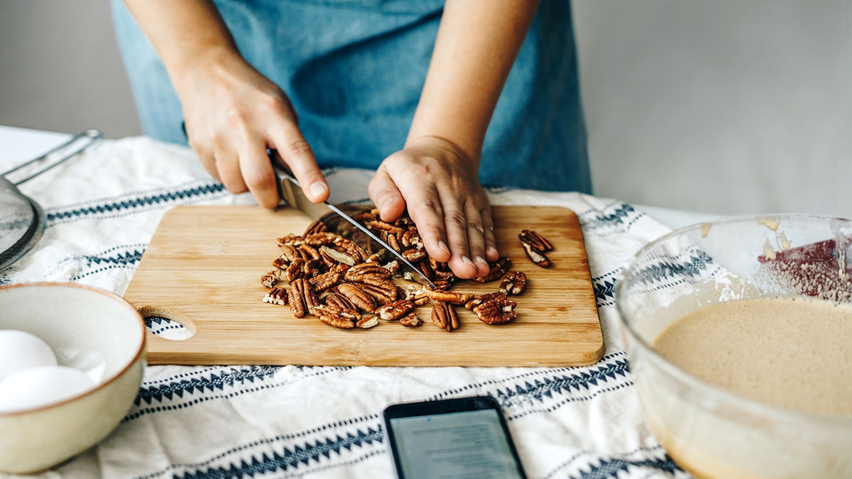 Person chopping pecans on a wooden cutting board while preparing baking ingredients in a home kitchen.
