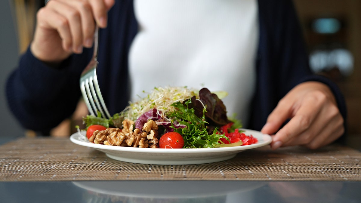 Person eating a fresh walnut and mixed greens salad with cherry tomatoes on a white plate.