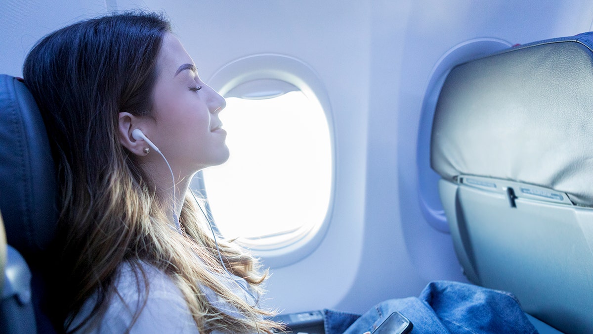 Woman relaxing with earphones and smartphone while seated by an airplane window during a flight.
