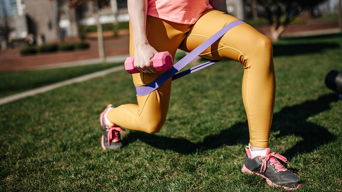 Woman performing a resistance band lunge with a dumbbell during an outdoor lower body workout on grass.