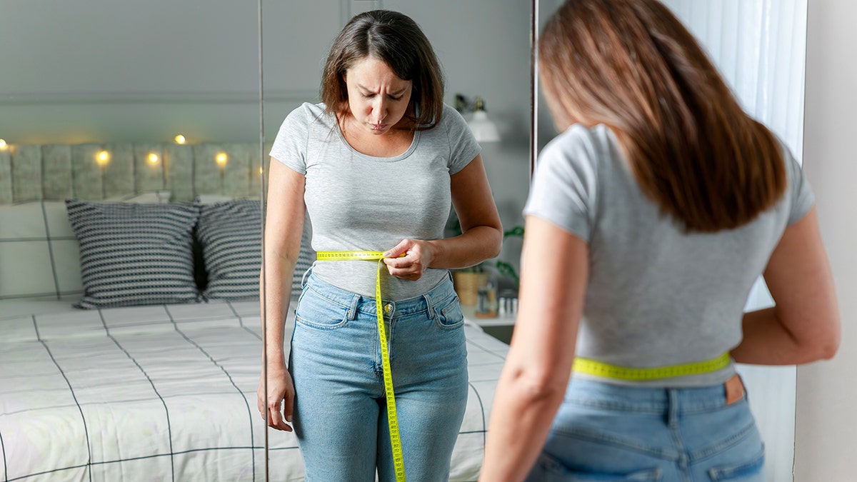 woman measures waist with tape measure in front of a mirror