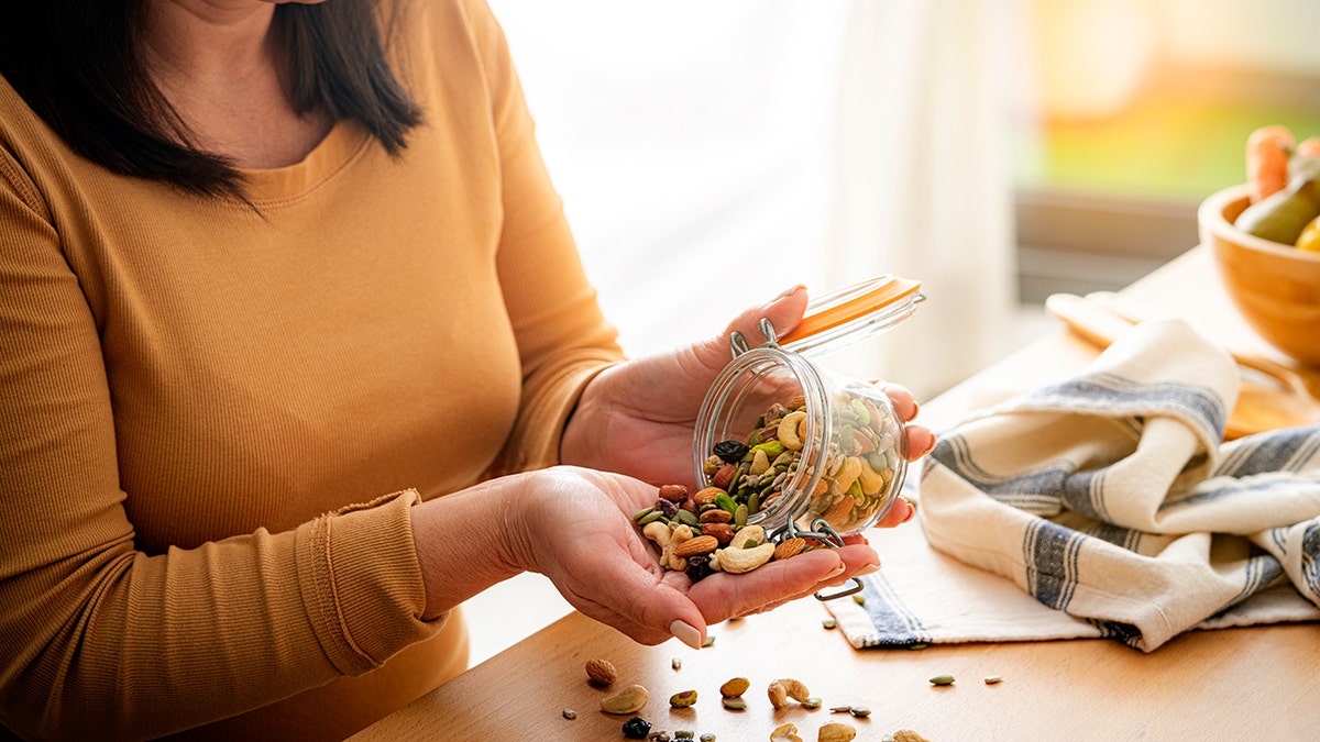 Person pouring mixed nuts and seeds from a glass jar into their hand in a bright kitchen setting.