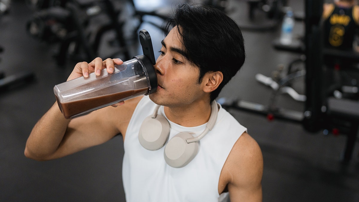 Young man sitting on bench at gym drinking protein shake with headphones around neck.