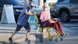 Andy Dick sits in a wheelchair as a man pushes him across a street