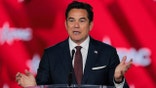 Dean Cain stands behind a clear podium with a serious expression, gesturing with both hands open as he speaks into a microphone against a red CPAC backdrop.