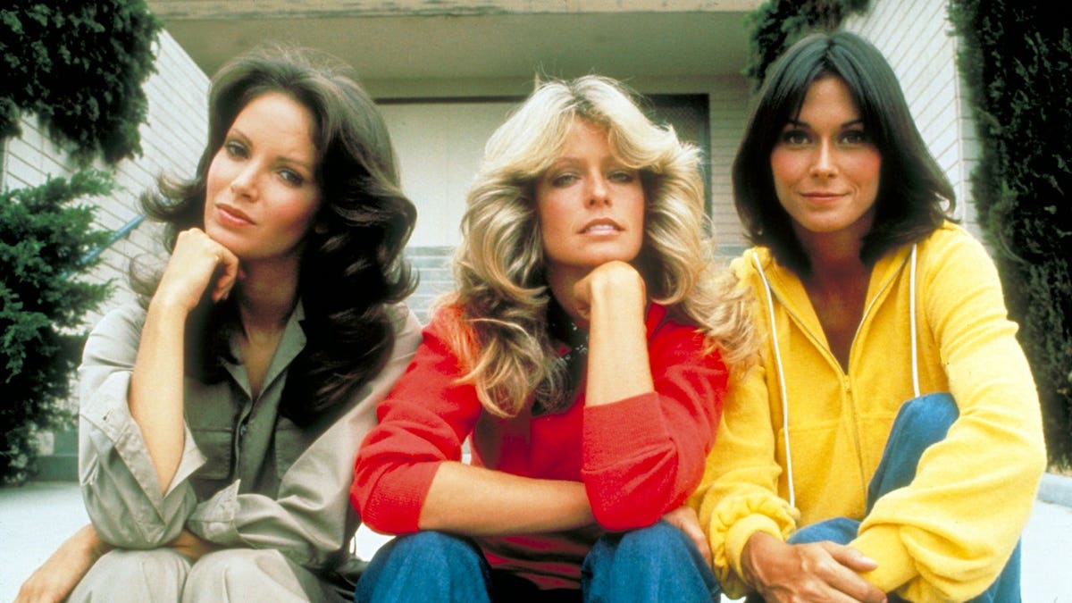 Jaclyn Smith, Farrah Fawcett and Kate Jackson standing together while looking toward the camera.