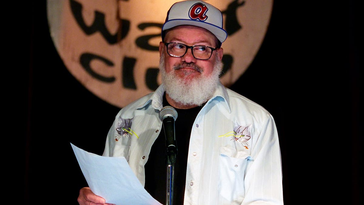 David Cross holds a piece of paper on a dimly lit stage.