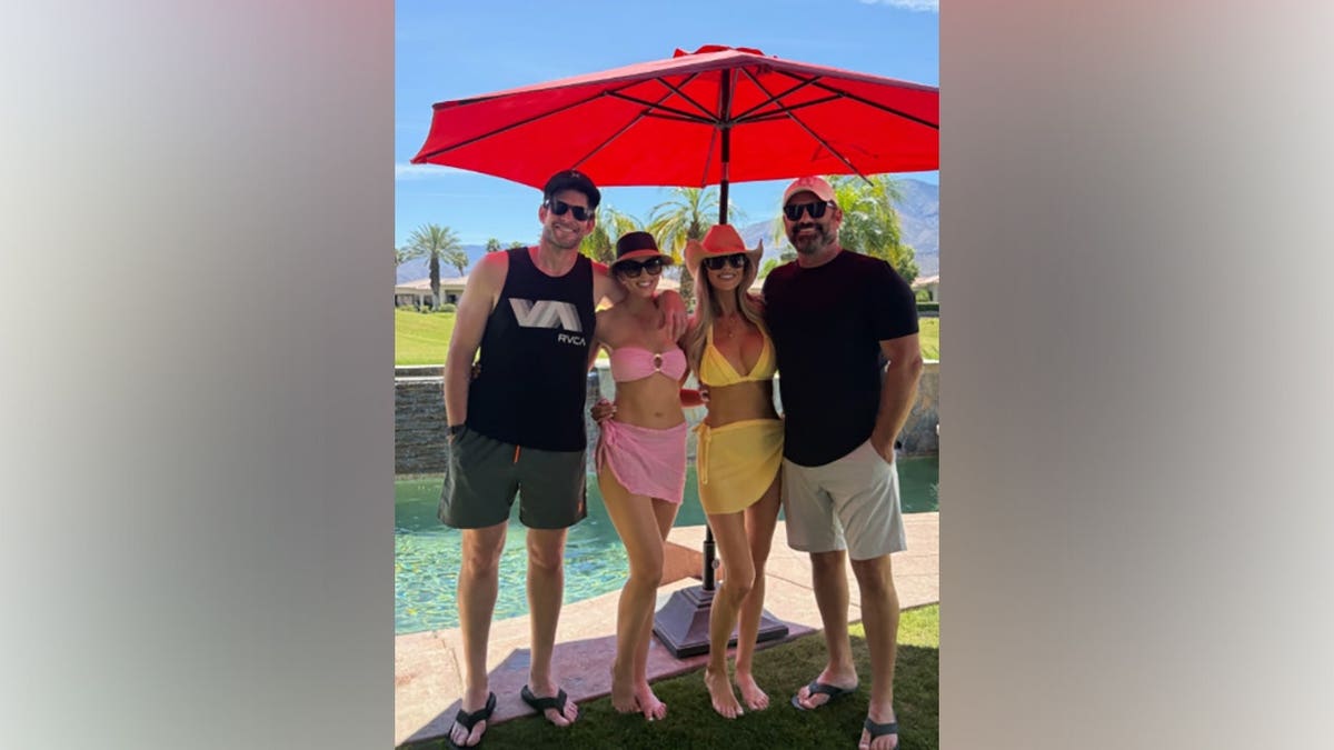 Tarek El Moussa, Heather Rae El Moussa, Christina Haack, and Christopher Larocca posing together by a pool under a red umbrella.