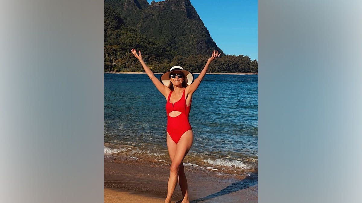 Jane Seymour wearing a red one-piece swimsuit and sun hat walking on a beach with mountains and ocean in the background