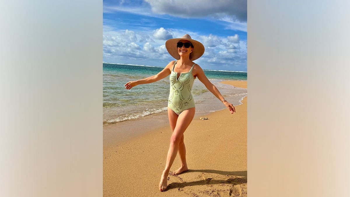 Jane Seymour walking along the shoreline in a green one-piece swimsuit with a sun hat and sunglasses under a bright blue sky
