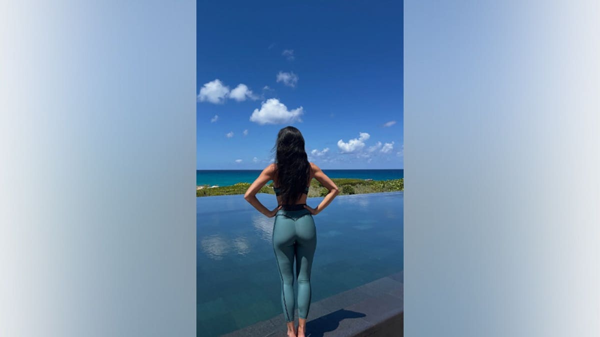 Nicole Scherzinger seen from behind with her hands on her hips, wearing teal leggings and looking at a tropical ocean view.