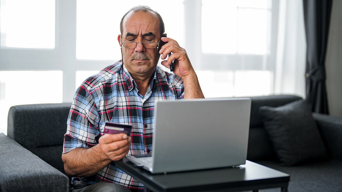 A man looks at his credit card while on the phone in front of a laptop.
