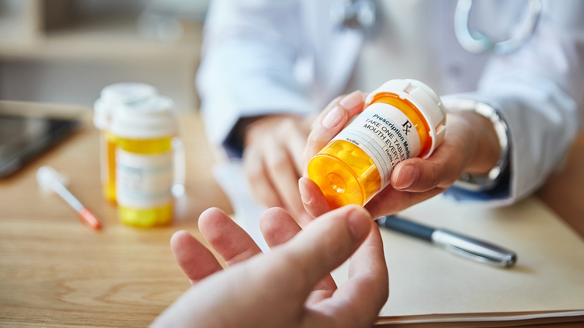 A person hands another person an orange prescription pill bottle.