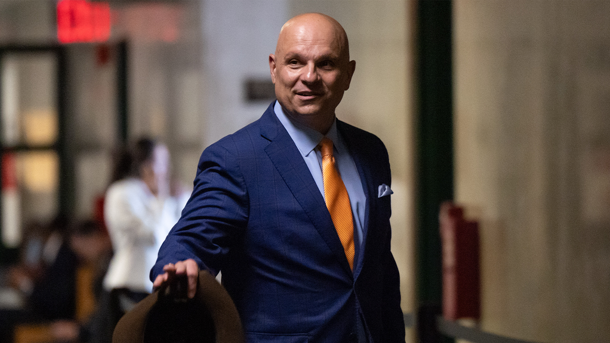 Attorney Arthur Aidala is holding a fedora in one hand as he gestures at someone out of frame. He is wearing a blue suit and shirt with an orange tie.