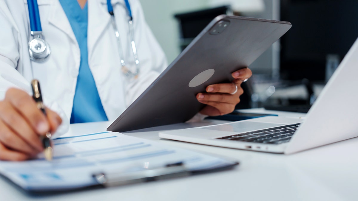 A doctor looks at a tablet and writes on a clipboard.