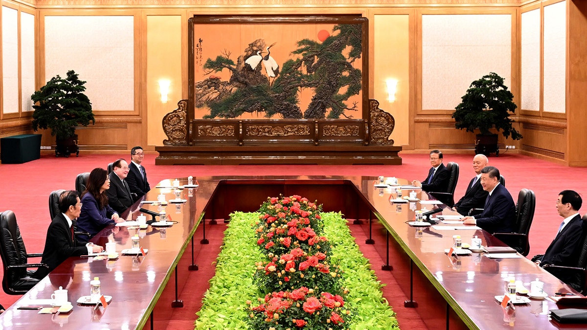 Los dignatarios se sientan en dos mesas situadas una frente a otra, con un tapiz al fondo y arreglos florales entre ambas mesas.