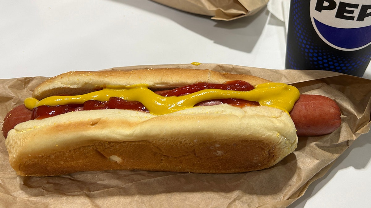 Costco hot dog seen with Pepsi soda cup and slathered in mustard and ketchup.