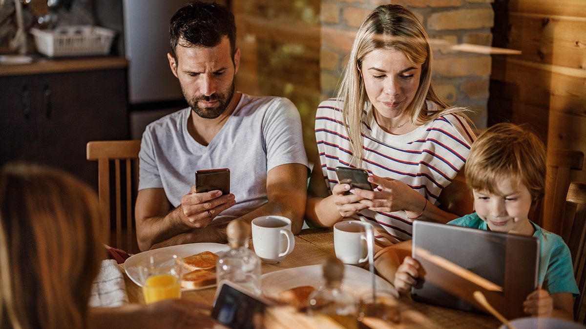 Young parents and their small kids using phones and tablets during morning time at dining table. The view is through glass.