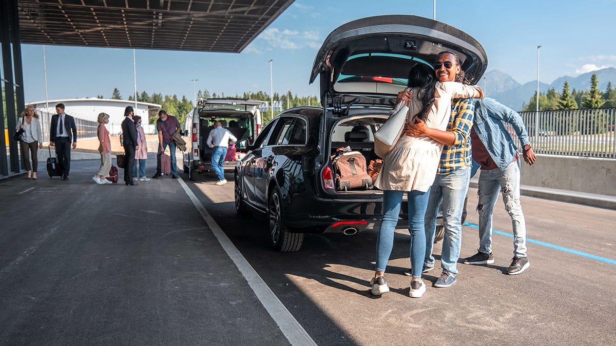 Diverse group of friends embracing and saying goodbye next to a car at an airport departure area
