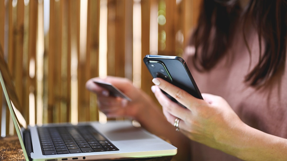 A woman holds her phone and a credit card in front of a laptop.