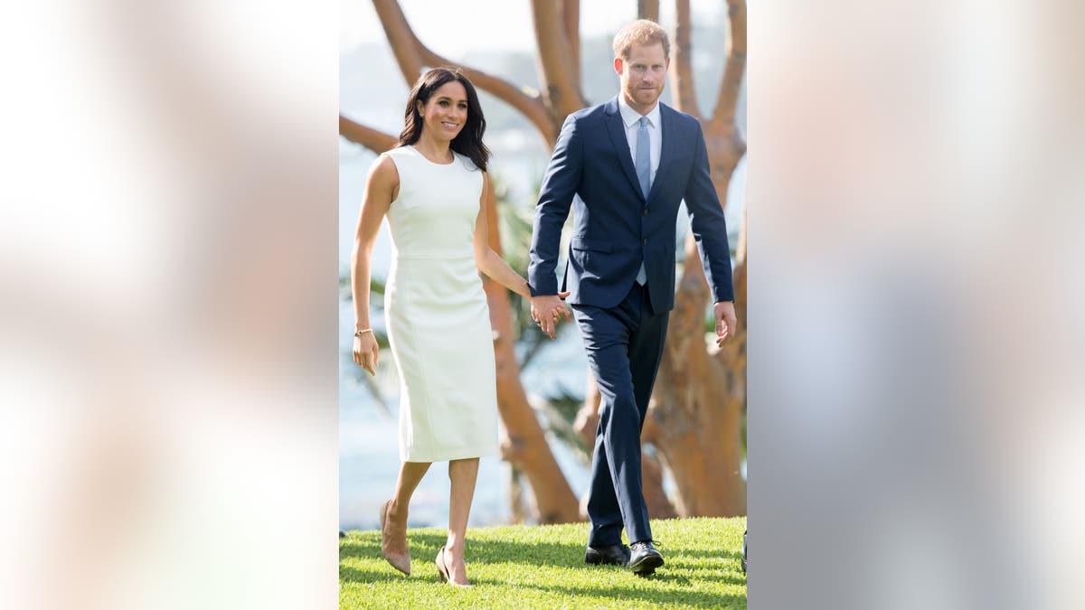 Prince Harry and Meghan Markle stand together outdoors at Admiralty House in Sydney during their royal tour, dressed formally and smiling.