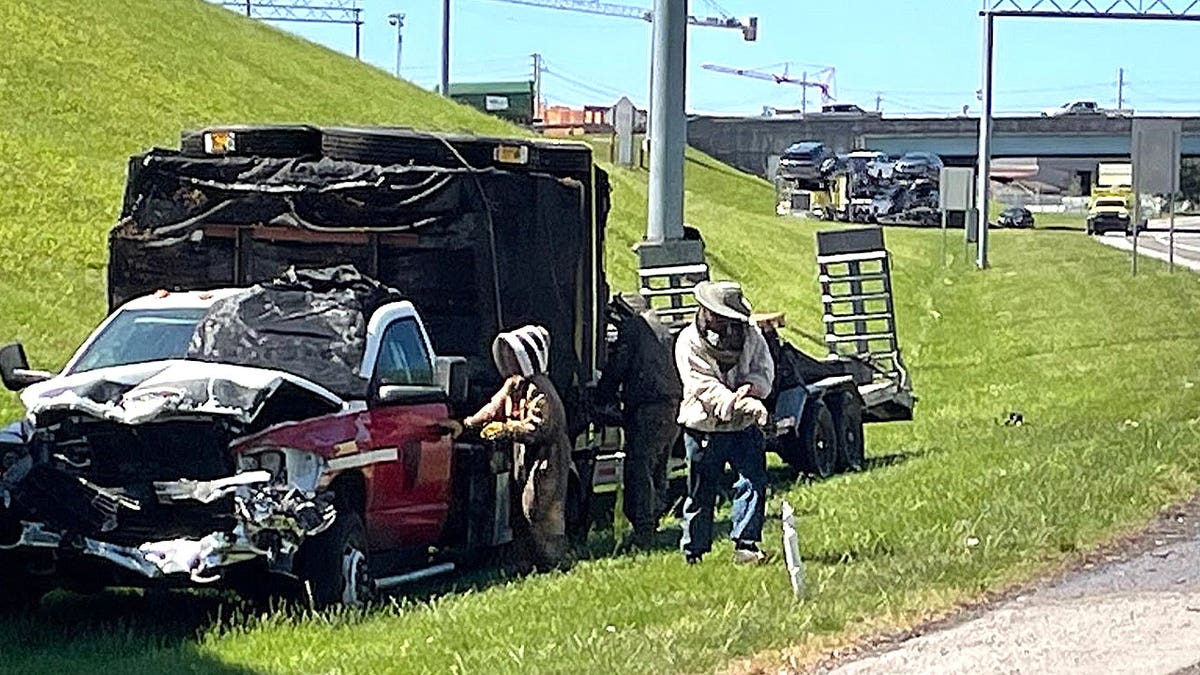 Several people wearing full-body protective beekeeping suits stand next to a wrecked truck