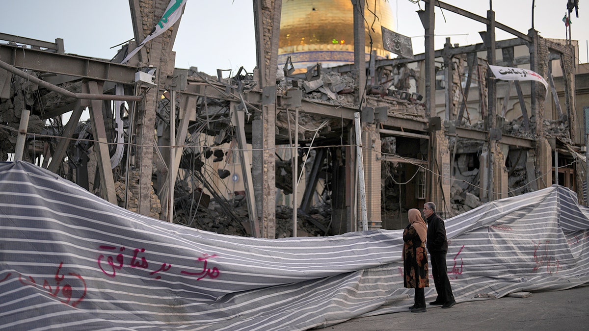 Pedestrians looking at a destroyed building with a mosque in the background.