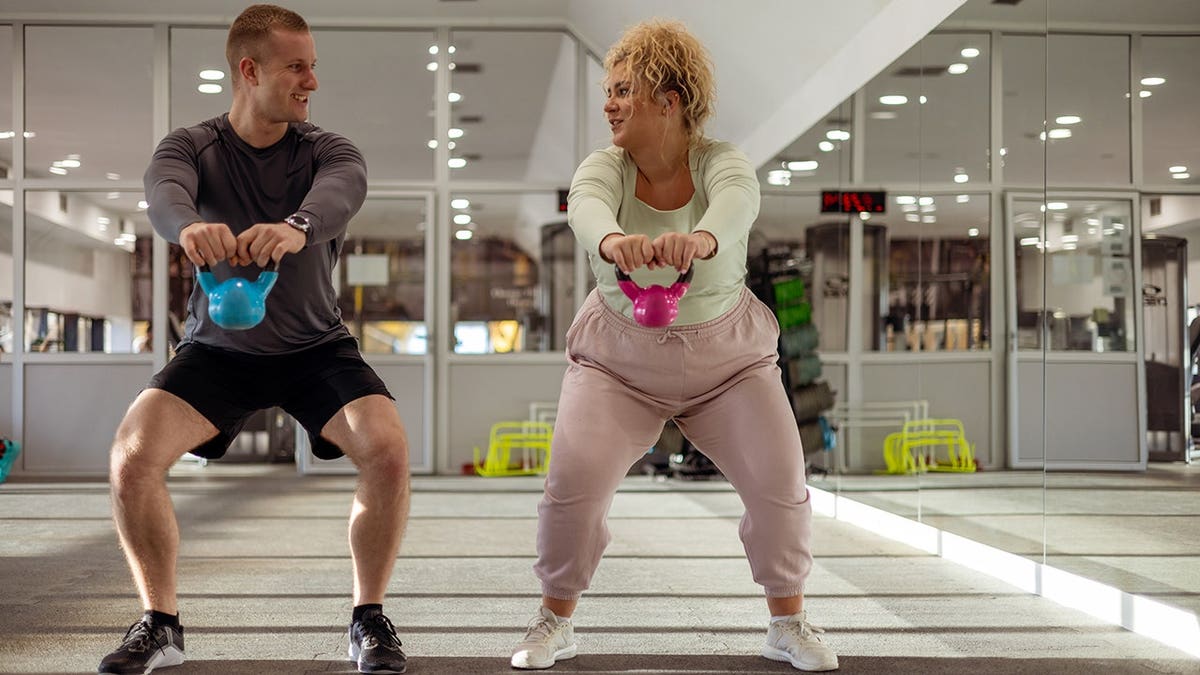Overweight female client lifting kettlebell with personal trainer in gym