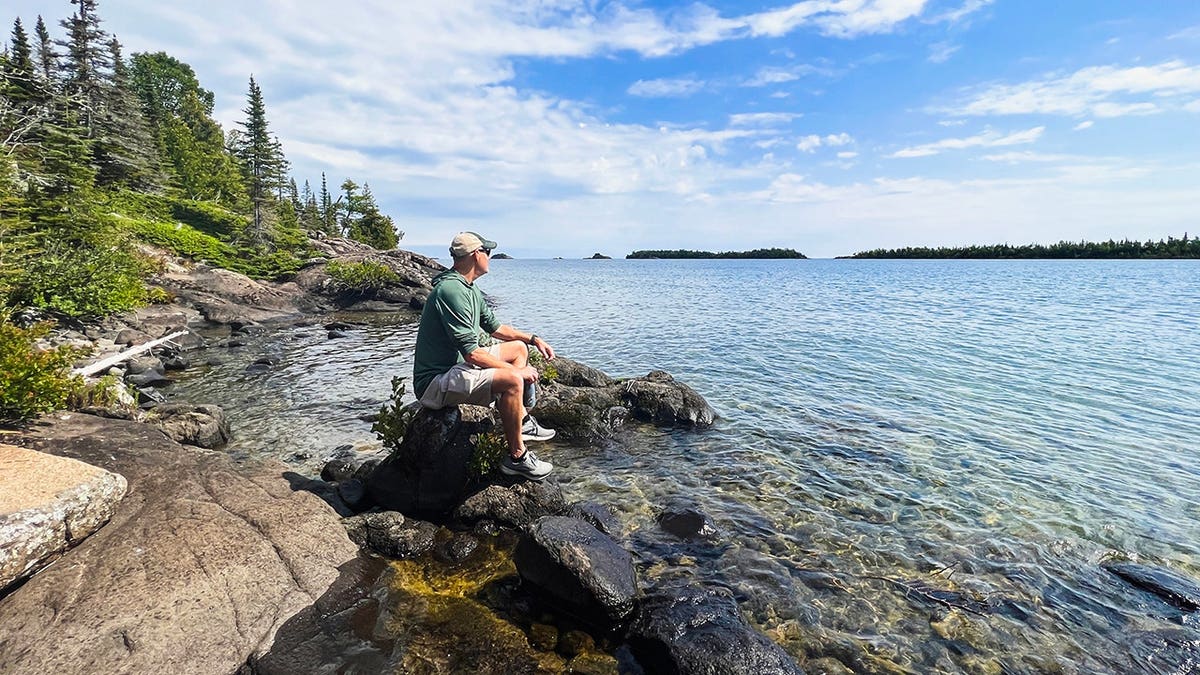 Um homem sentado em uma grande rocha com vista para o Lago Superior no Parque Nacional Isle Royale, em Michigan