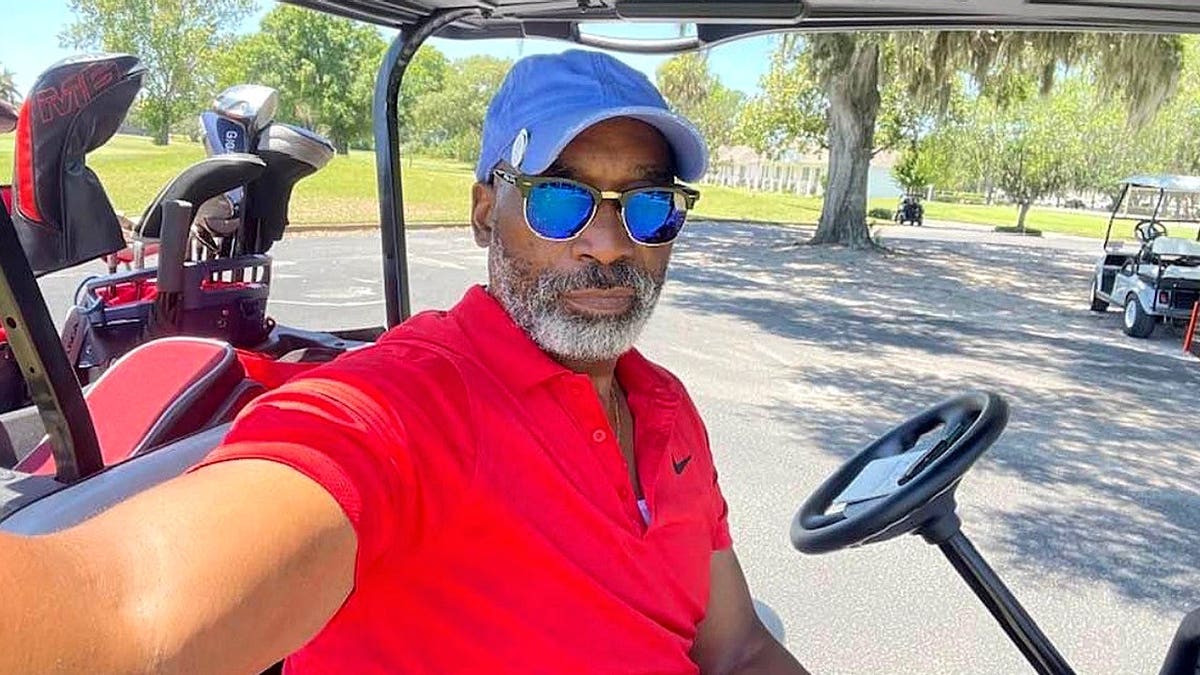 Leslie williams wearing red polo, blue hat and sunglasses, as he takes a selfie in a golfing cart