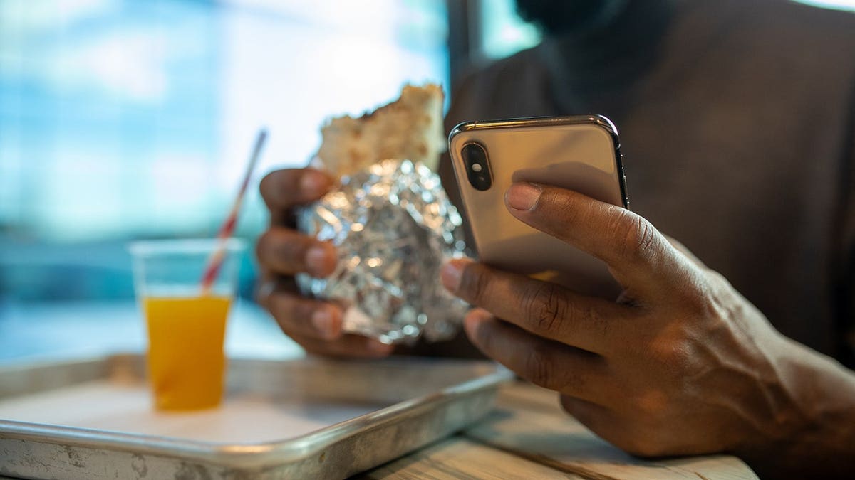 Adult man holding foil-wrapped burrito and scrolling on smartphone during meal.