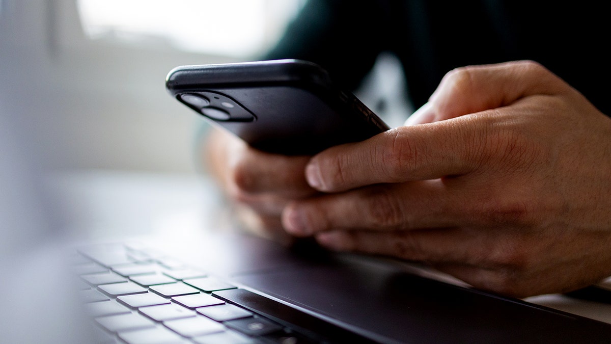 A person uses an iPhone next to a laptop.