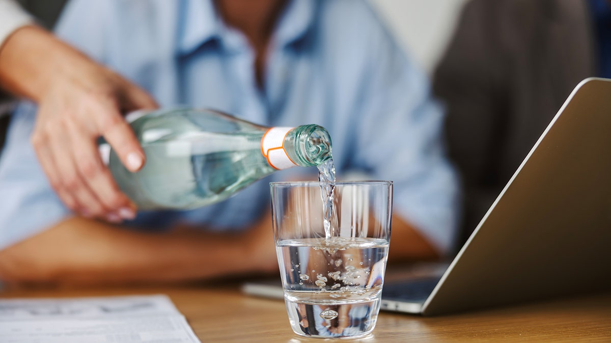Woman's hand pouring refreshing water from a glass bottle into a drinking glass on a wooden table, emphasizing healthy hydration during a busy workday or business meeting