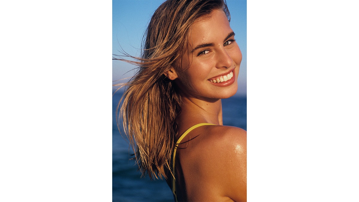 Model Niki Taylor glances over her shoulder with a slight grin while wearing a bright yellow swimsuit, with the ocean behind her.