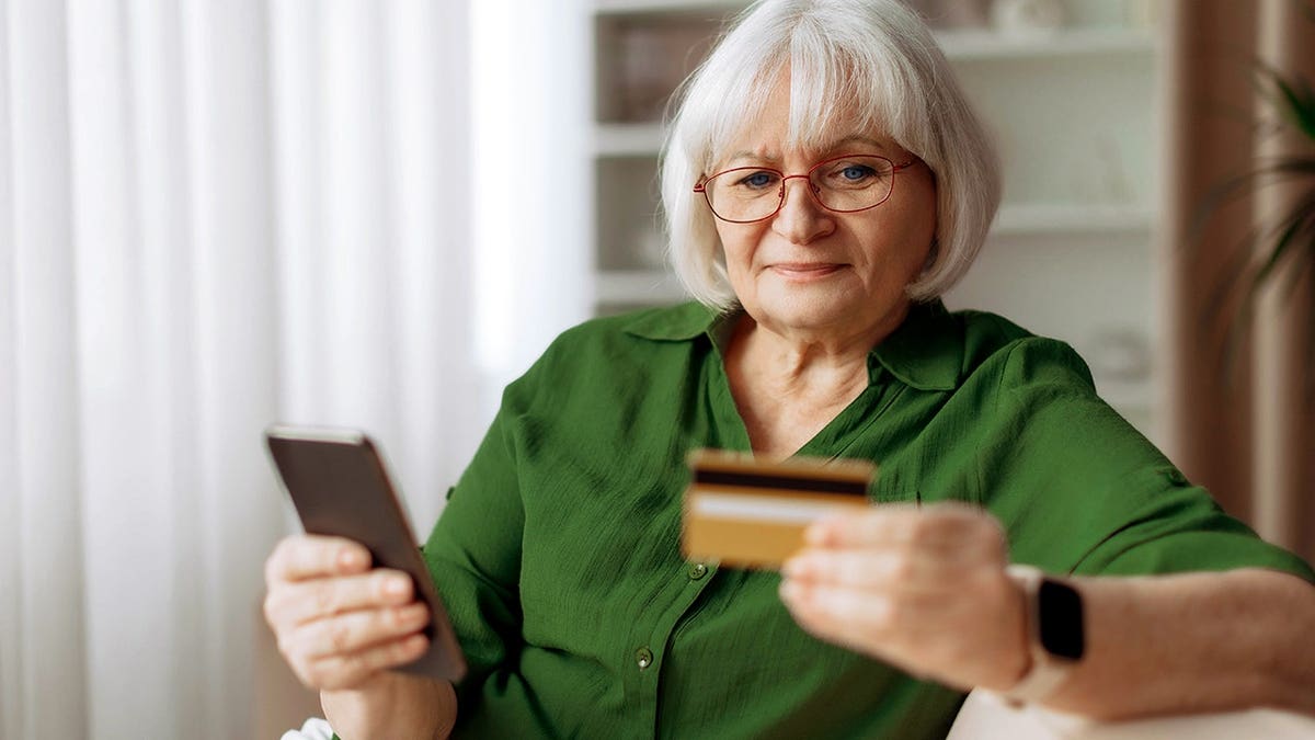 An elderly woman looks at her credit card while holding her phone.