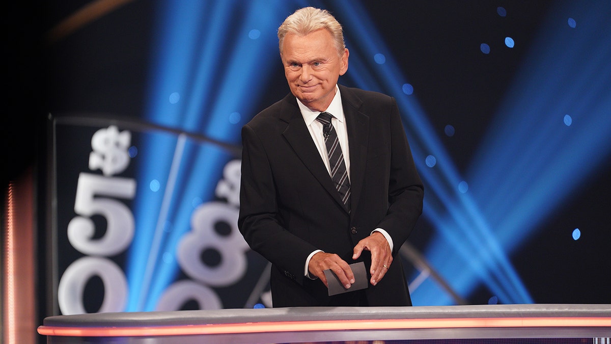 Pat Sajak stands on the set of Celebrity Wheel of Fortune, smiling as he hosts the game show.