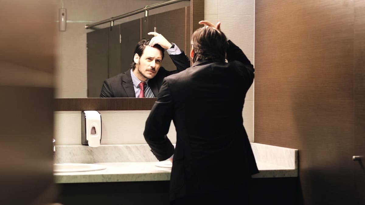 Businessman looking at receding hairline in office bathroom mirror